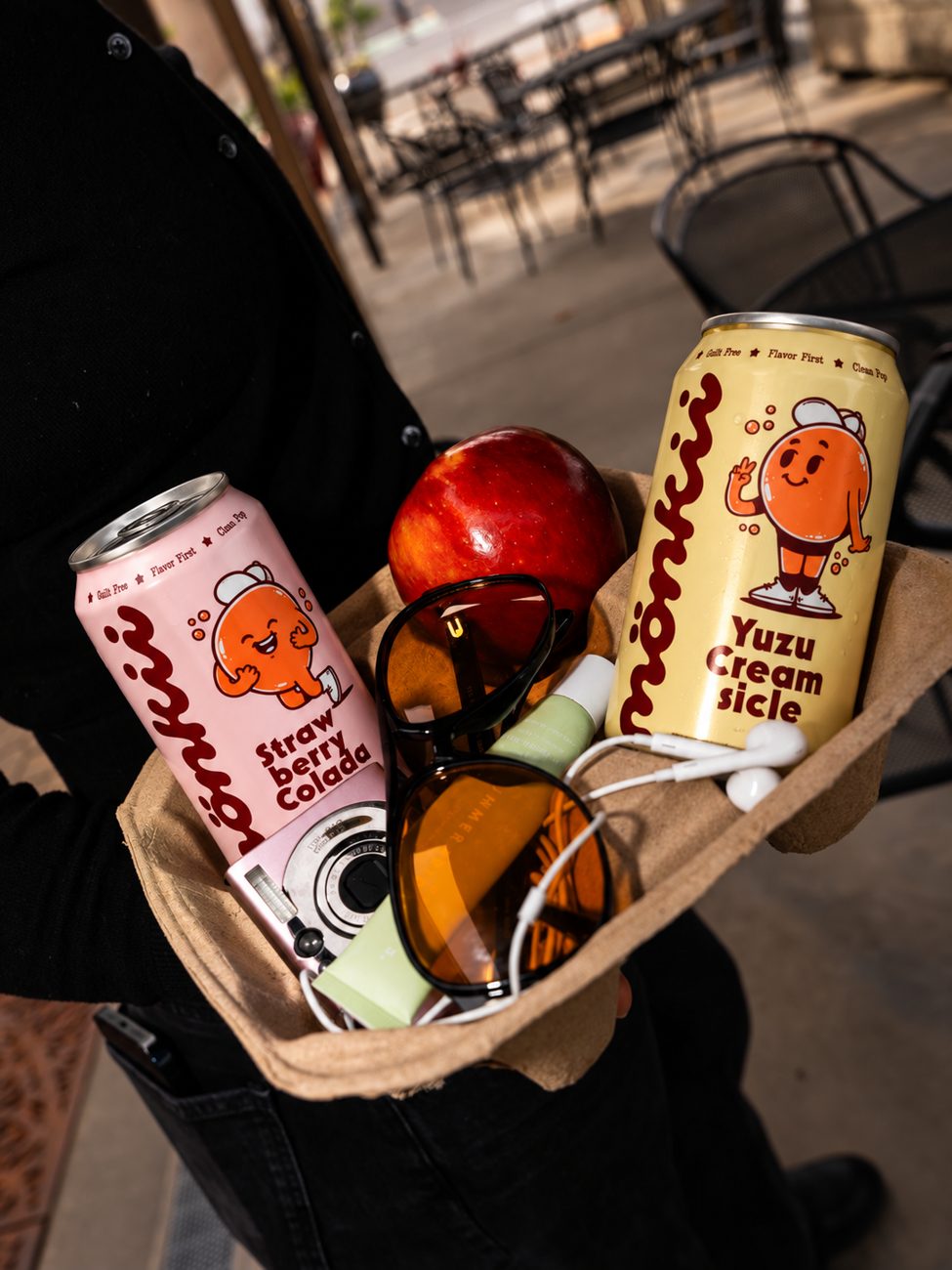 Strawberry Colada and Yuzu Creamsicle cans in a carry tray with an apple, sunglasses, a camera and earbuds